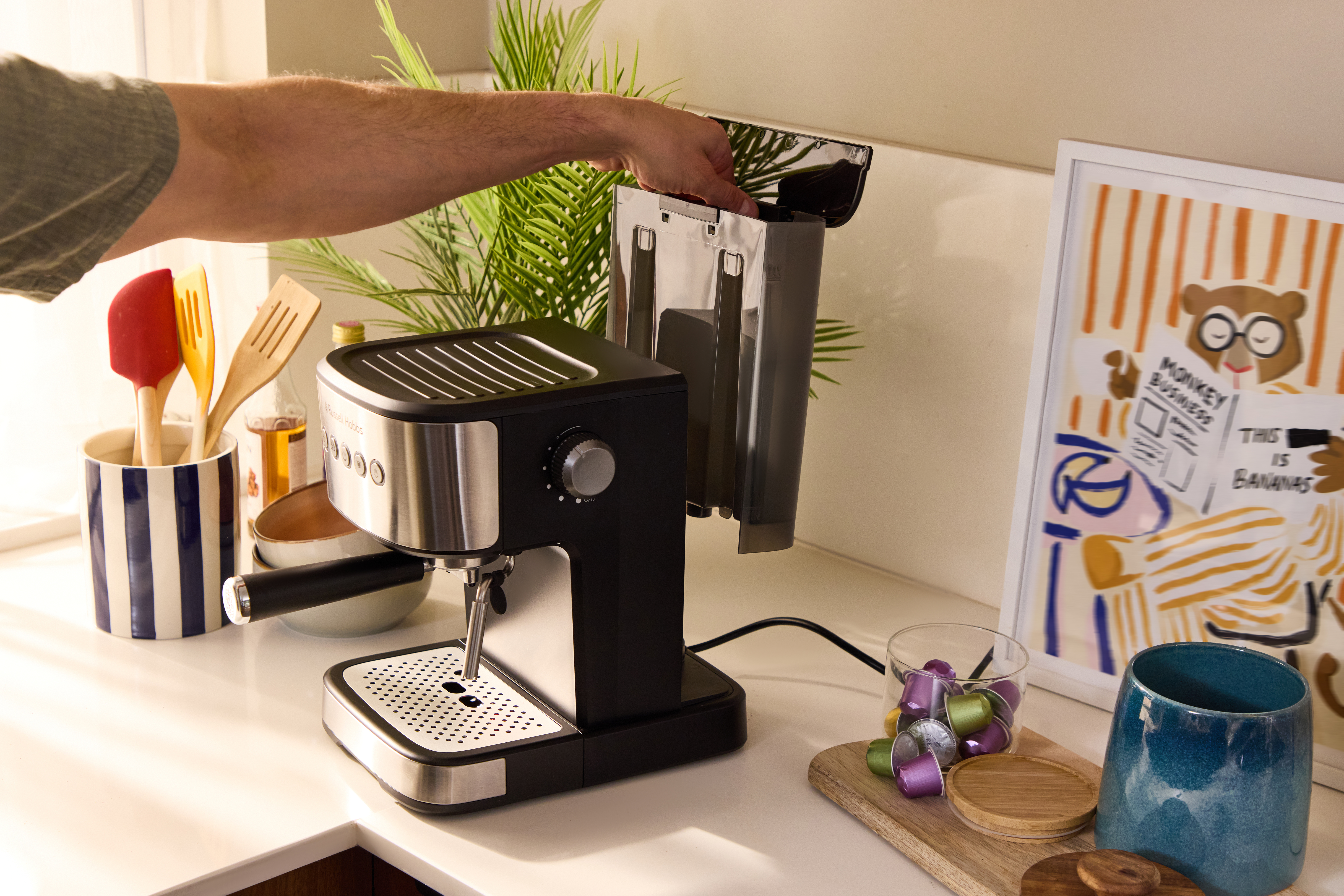 Eine Hand befüllt eine Kaffeemaschine mit Wasser. Daneben stehen Utensilien und eine Bild.