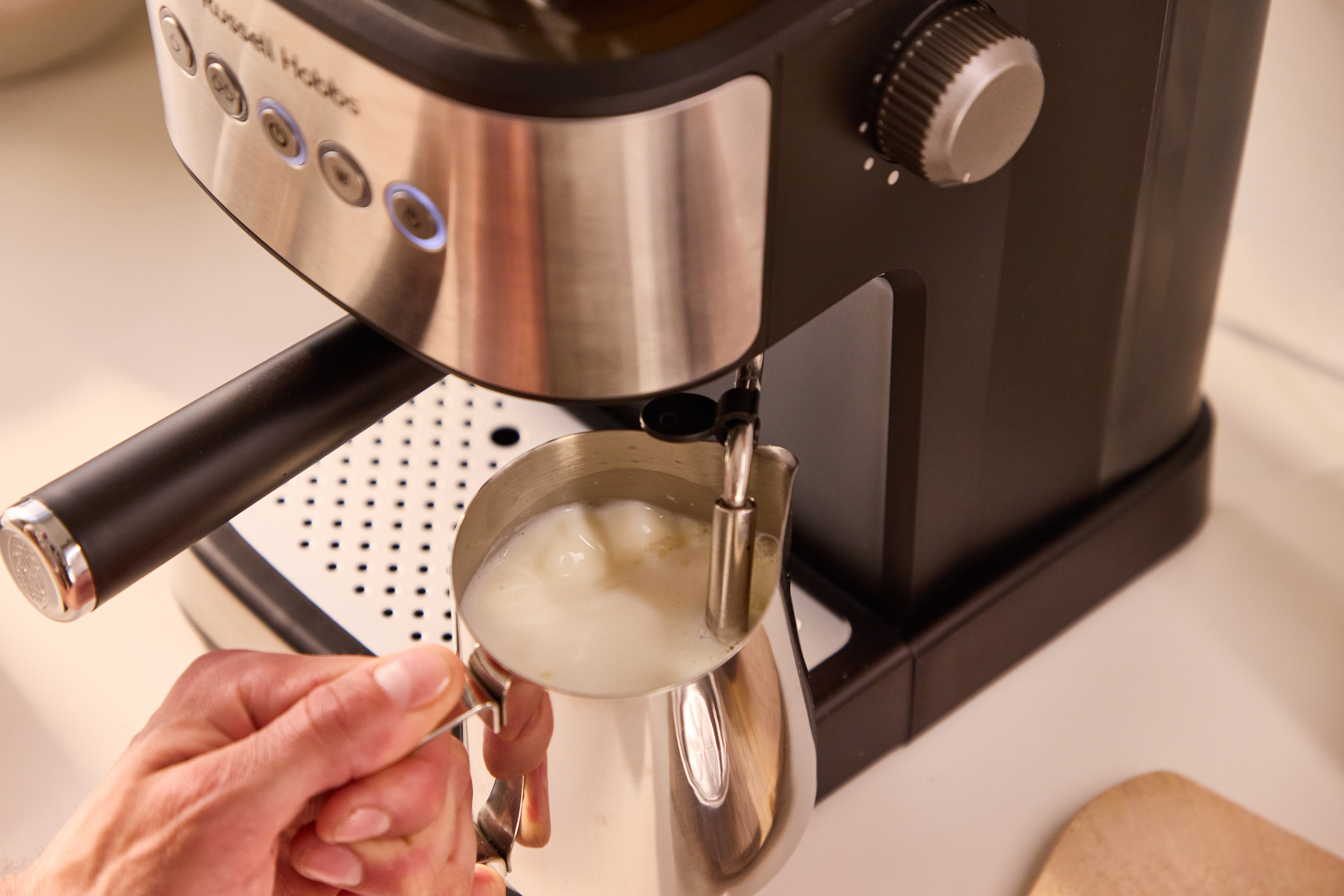 Eine Hand hält einen Milchaufschäumer mit aufgeschäumter Milch unter einer Kaffeemaschine.