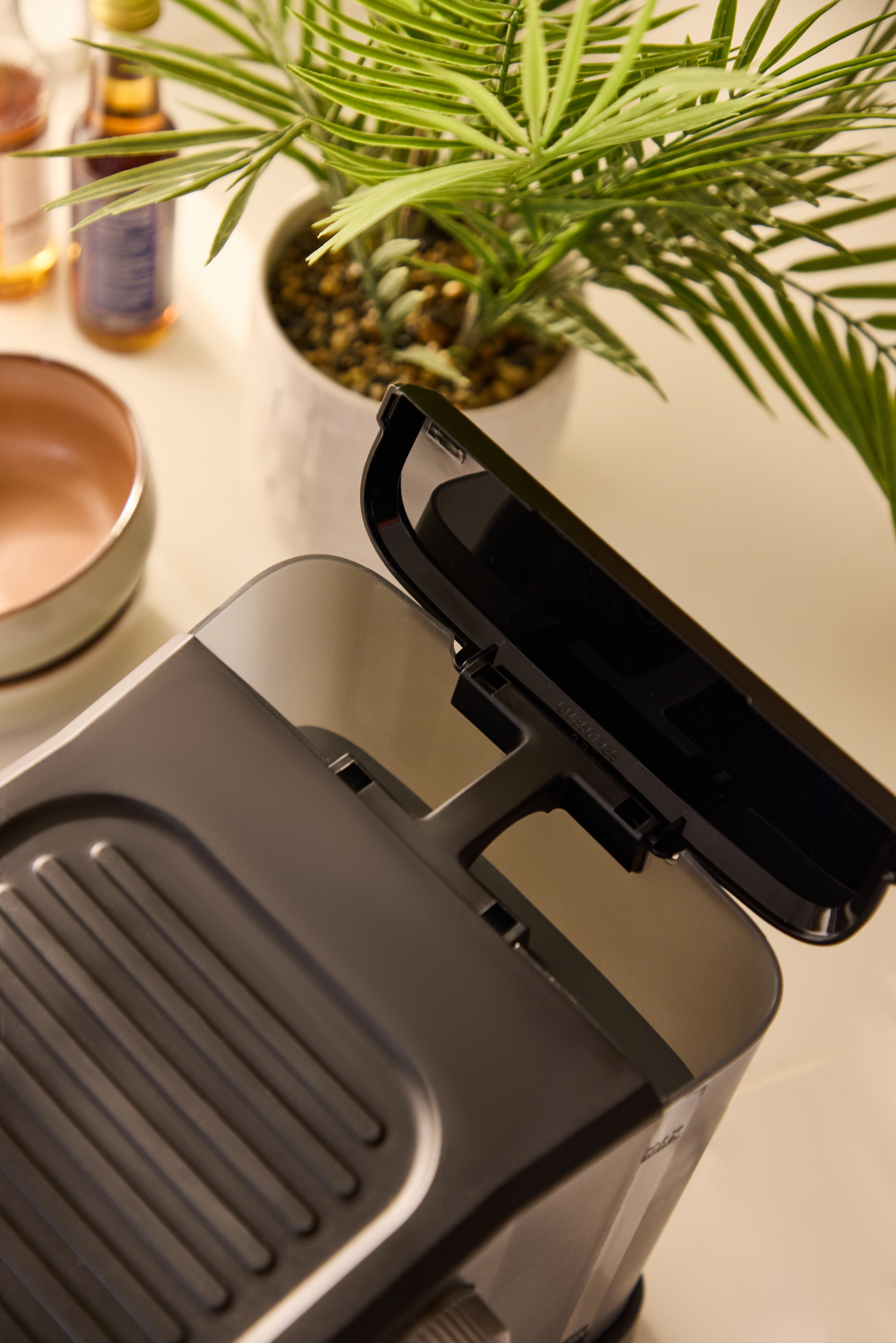 Eine Kaffeemaschine mit offenem Deckel, daneben eine Pflanze und Flaschen.