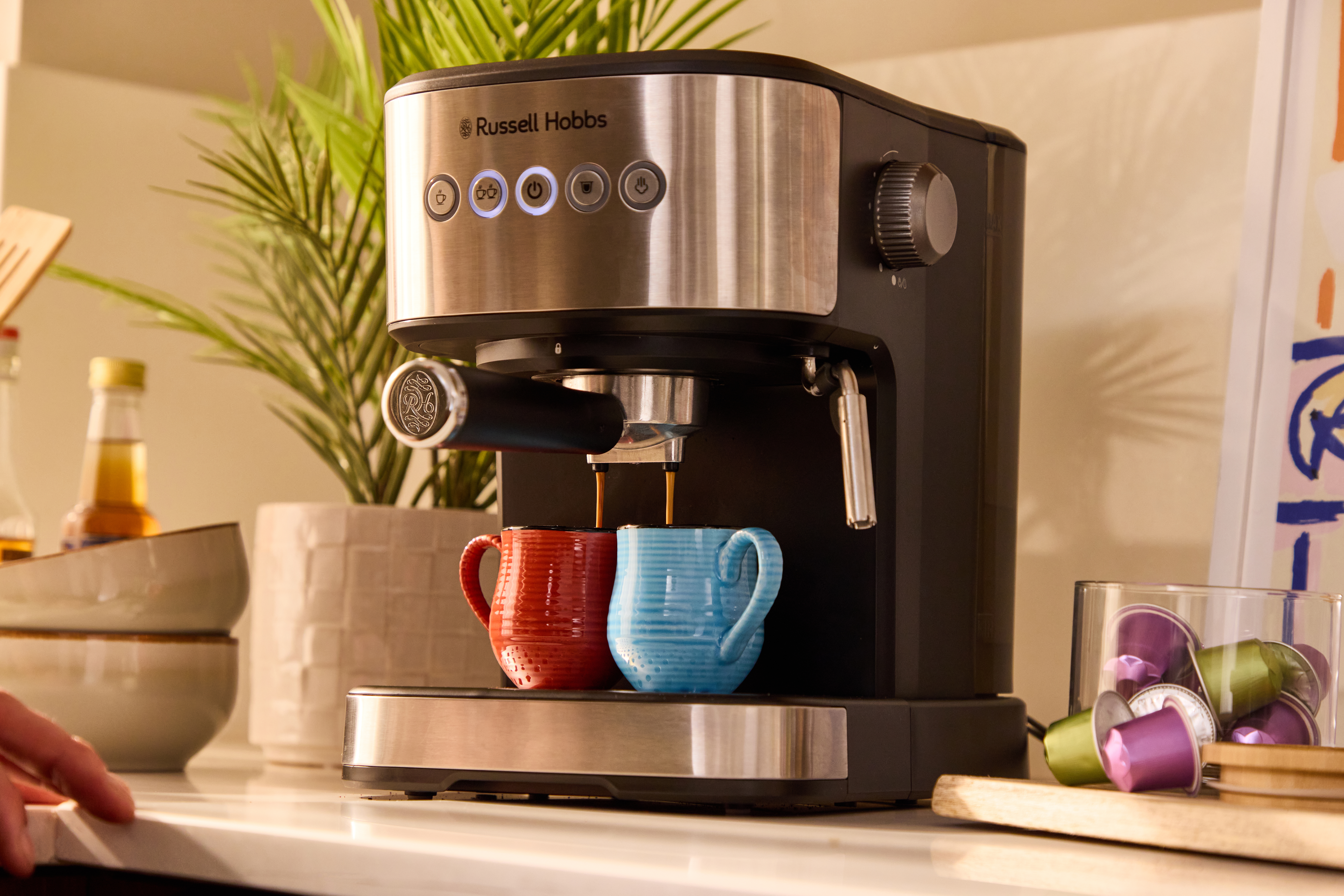 Eine Espressomaschine brüht Kaffee in zwei Tassen, rot und blau, daneben Kapseln.