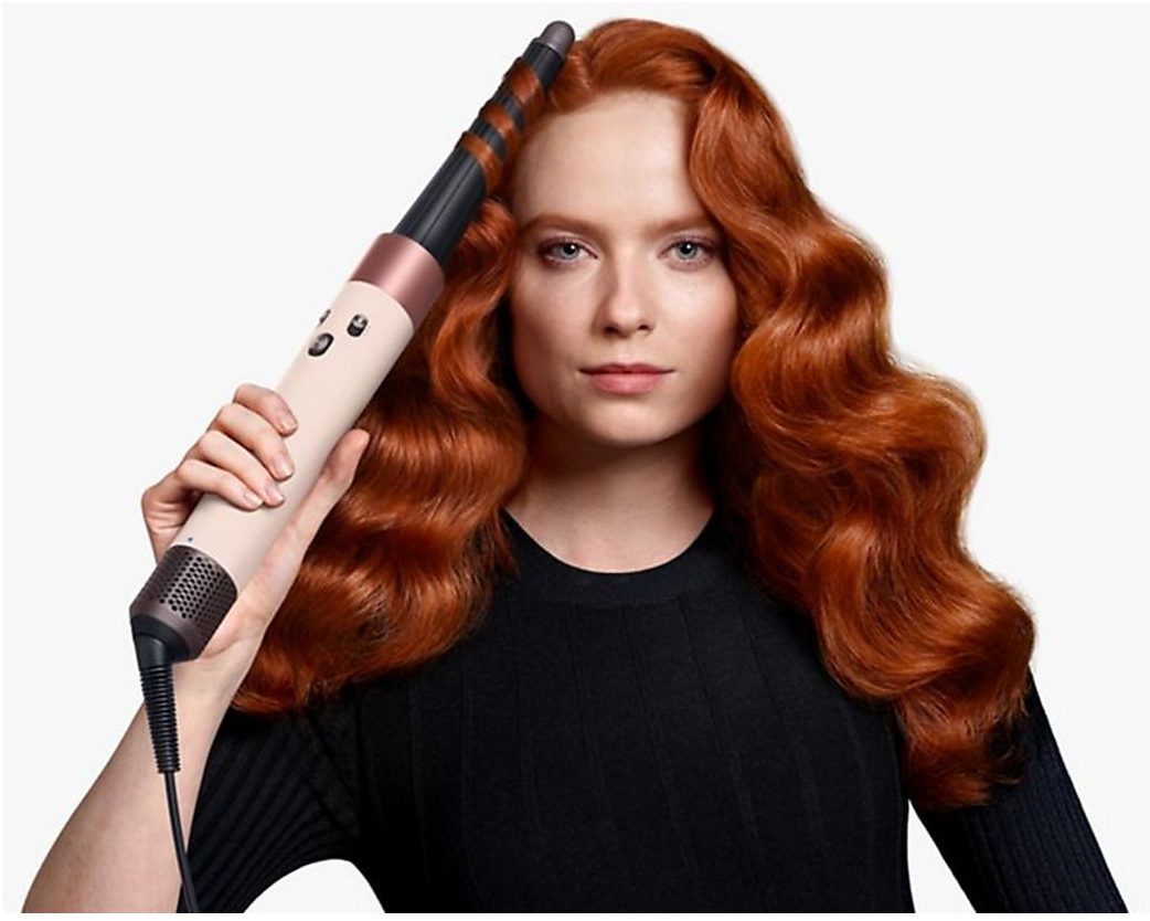 Femme aux cheveux roux tient un fer à friser avec un corps rose beige et des accents noirs.