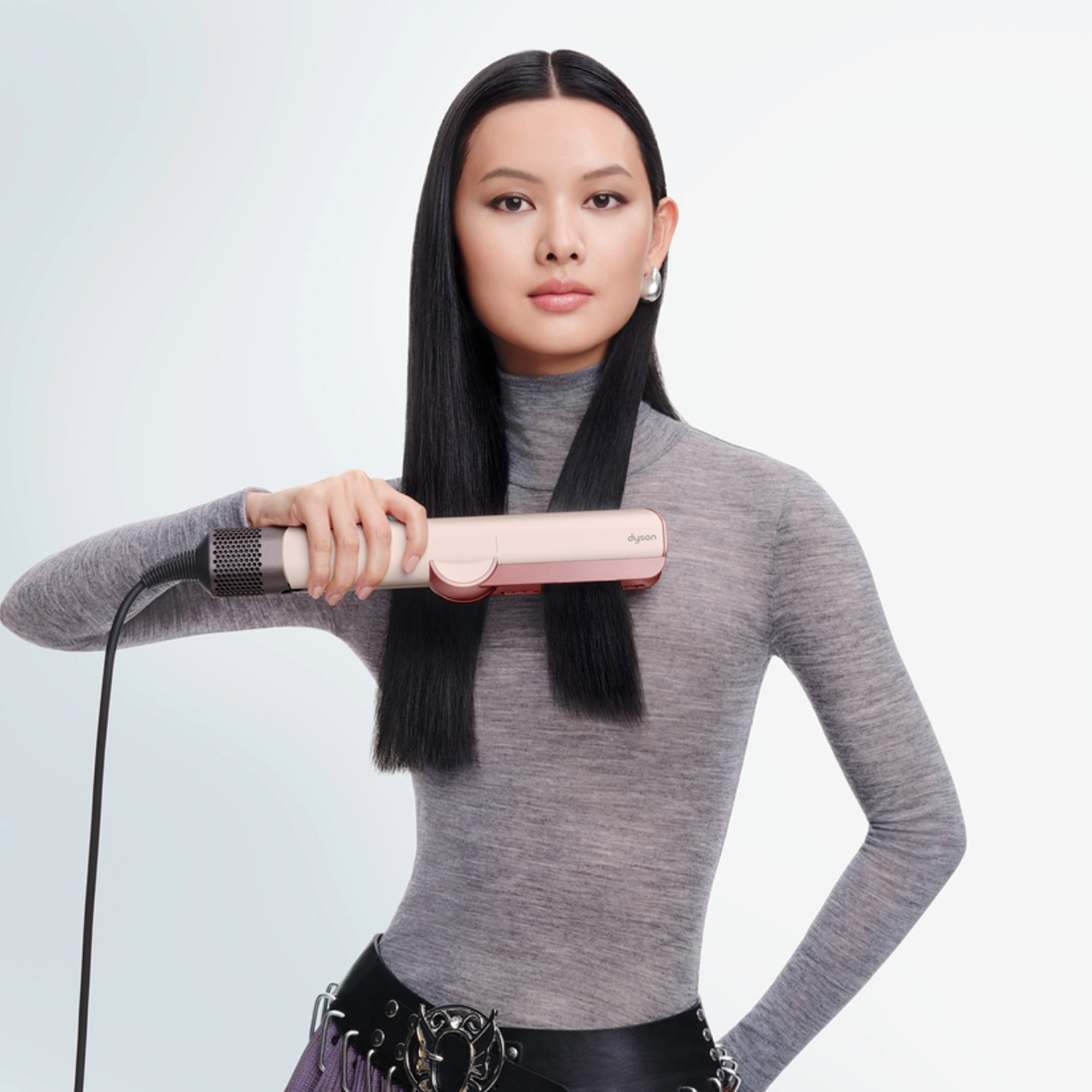 Une femme aux longs cheveux noirs les lisse avec un fer Dyson rose.