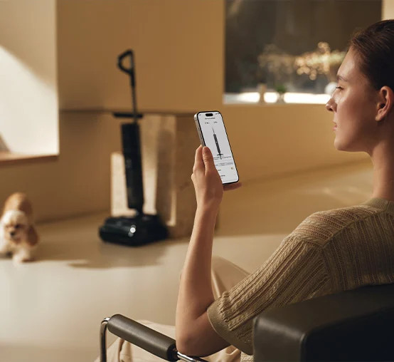 Kobieta trzyma telefon z aplikacją odkurzacza; pies i odkurzacz w tle.