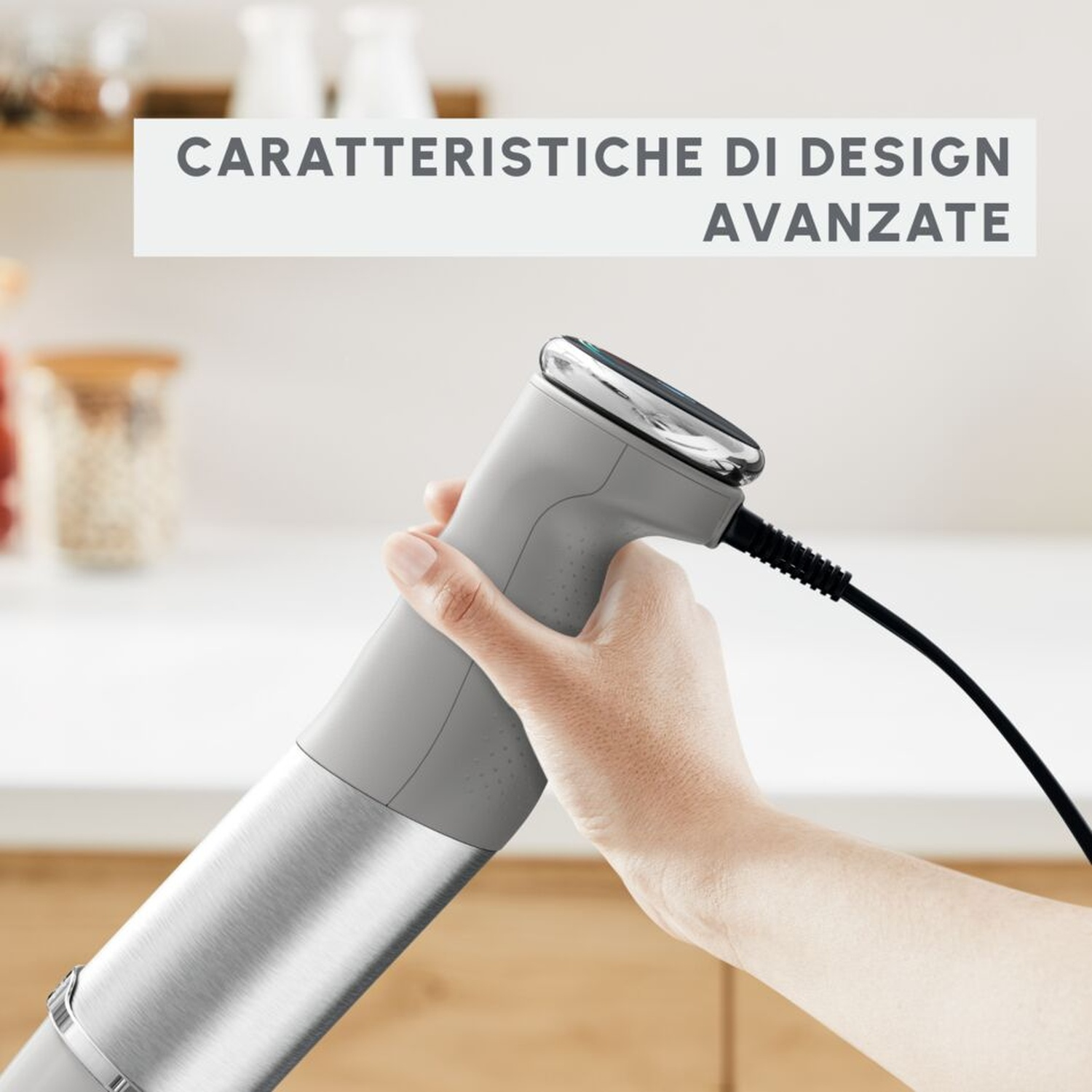 Una mano tiene un frullatore a immersione grigio con base in metallo argentato e cavo nero.