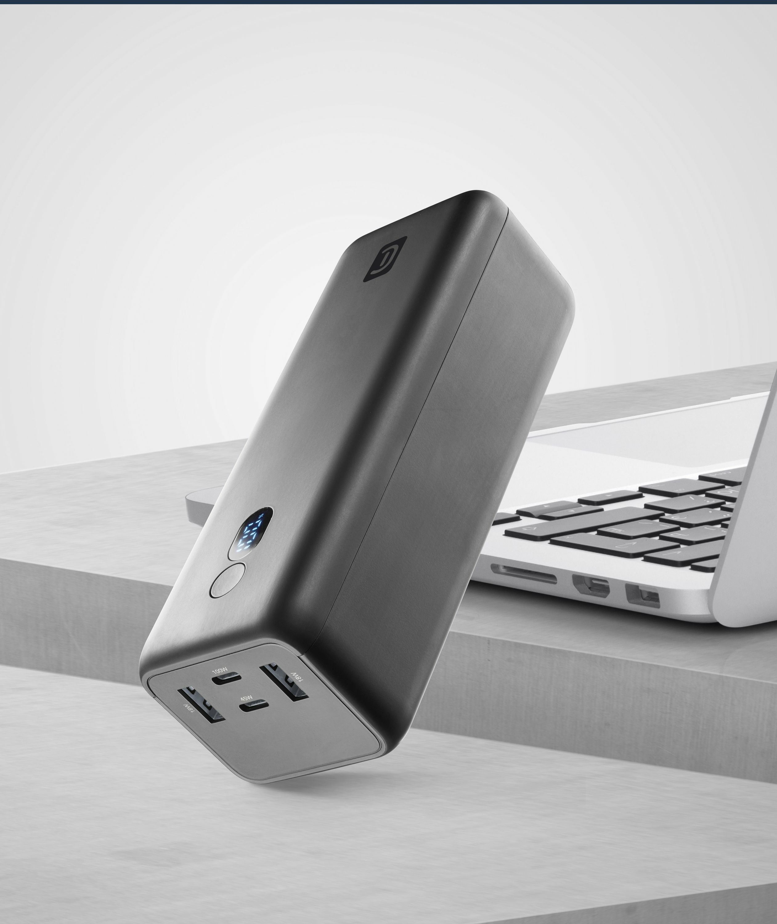 Un banco de energía negro con puertos USB junto a un portátil en una plataforma gris.