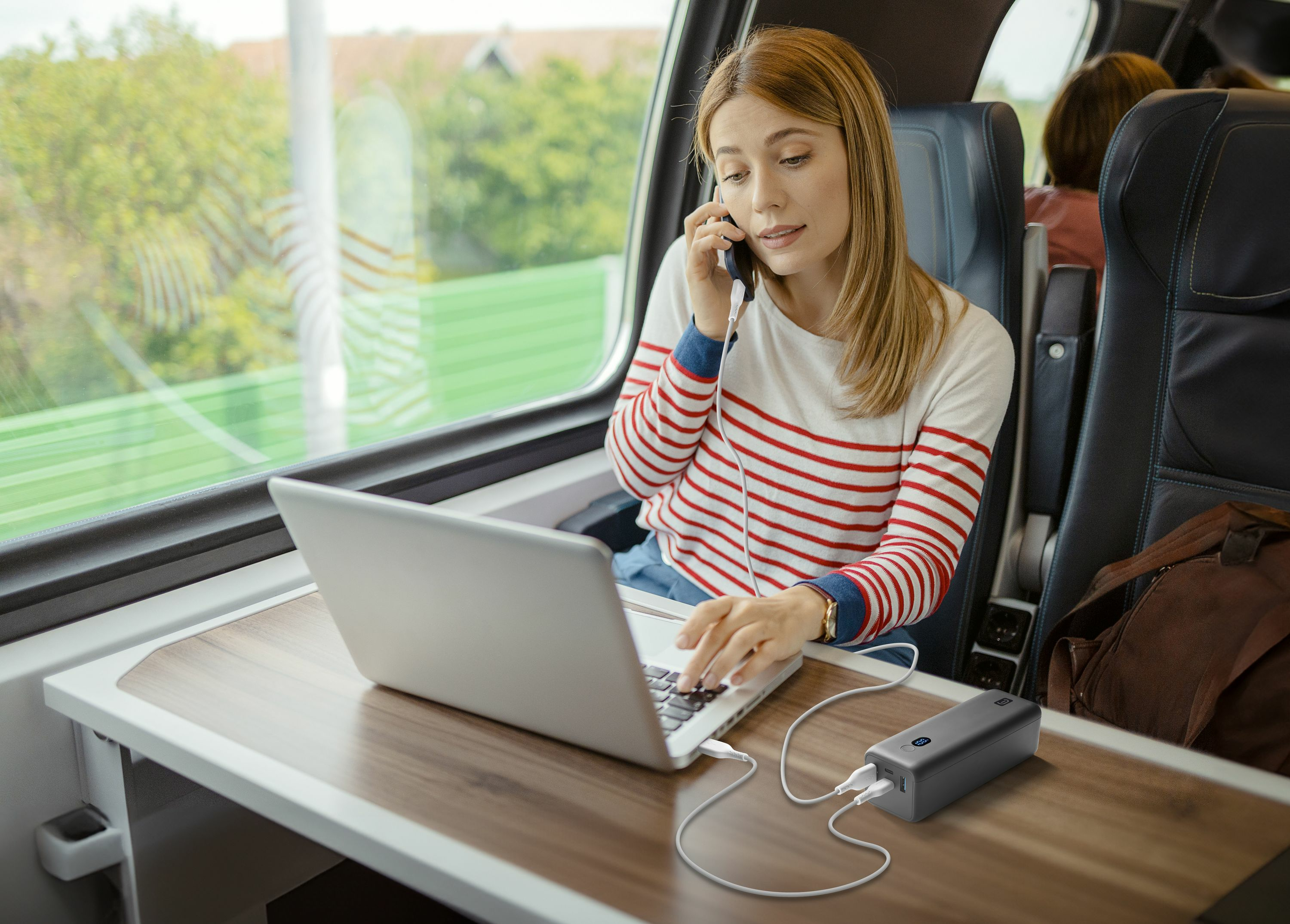 Mujer habla por teléfono, teclea en portátil, carga power bank en tren.