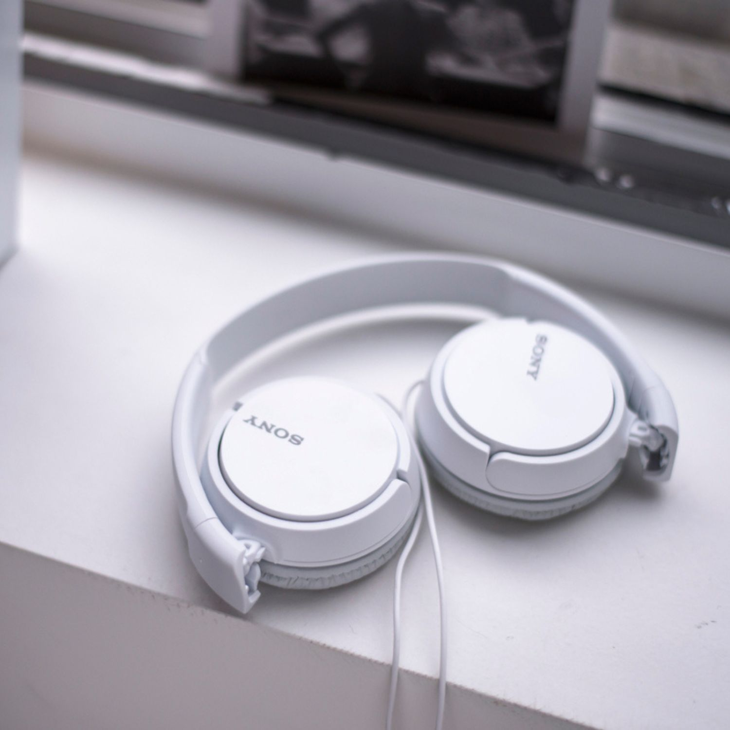 Auriculares Sony blancos plegados sobre superficie blanca, con libros al fondo.