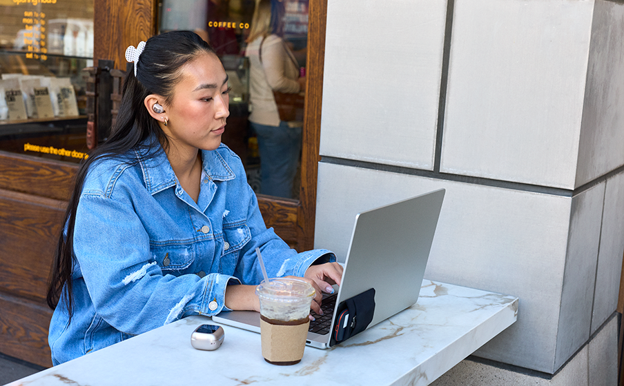 Eine junge Frau mit Kopfhörern arbeitet an einem Laptop im Freien, neben ihr ein Kaffee.