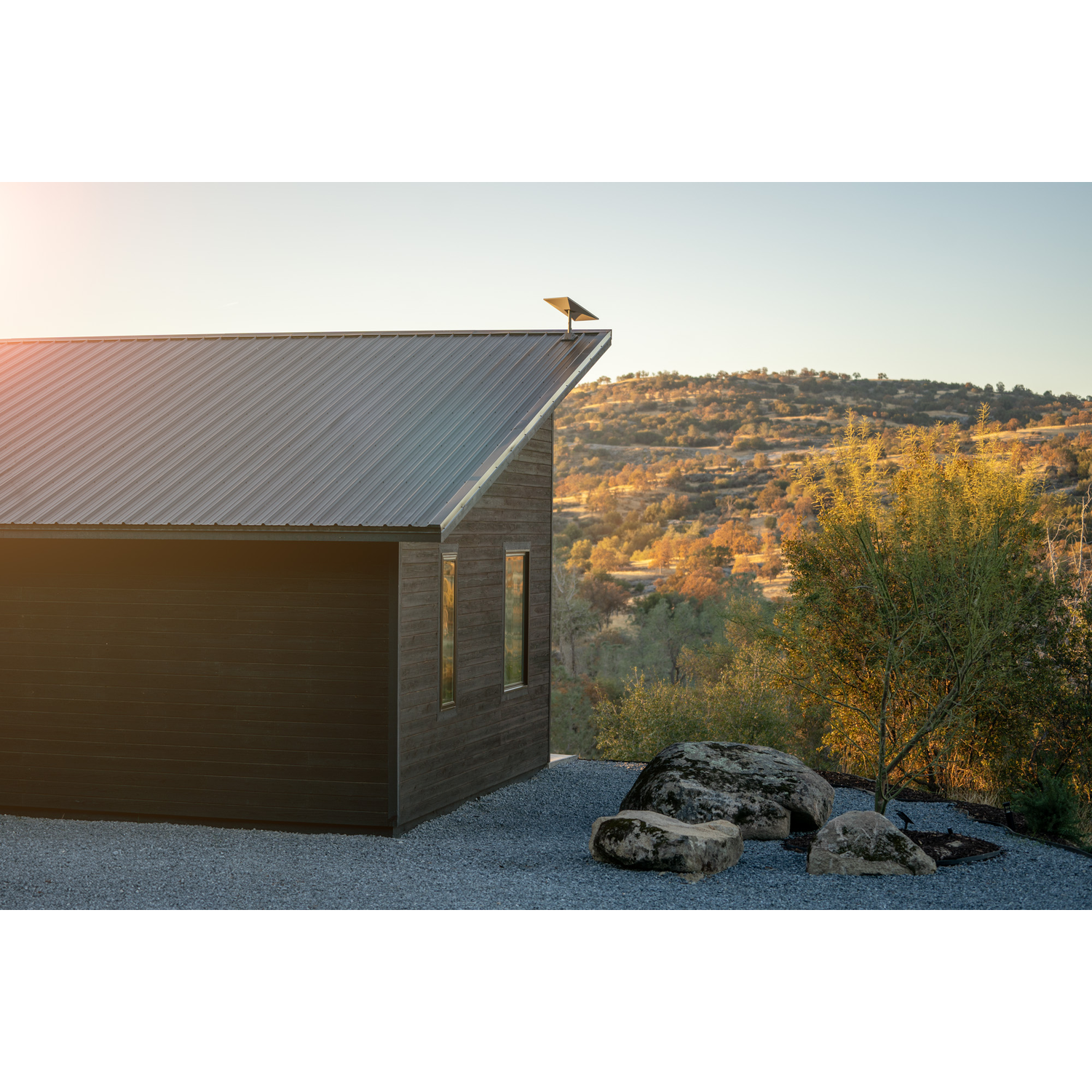 Modern donkerhouten huis met metalen dak in rotsachtig landschap, zonsondergang.