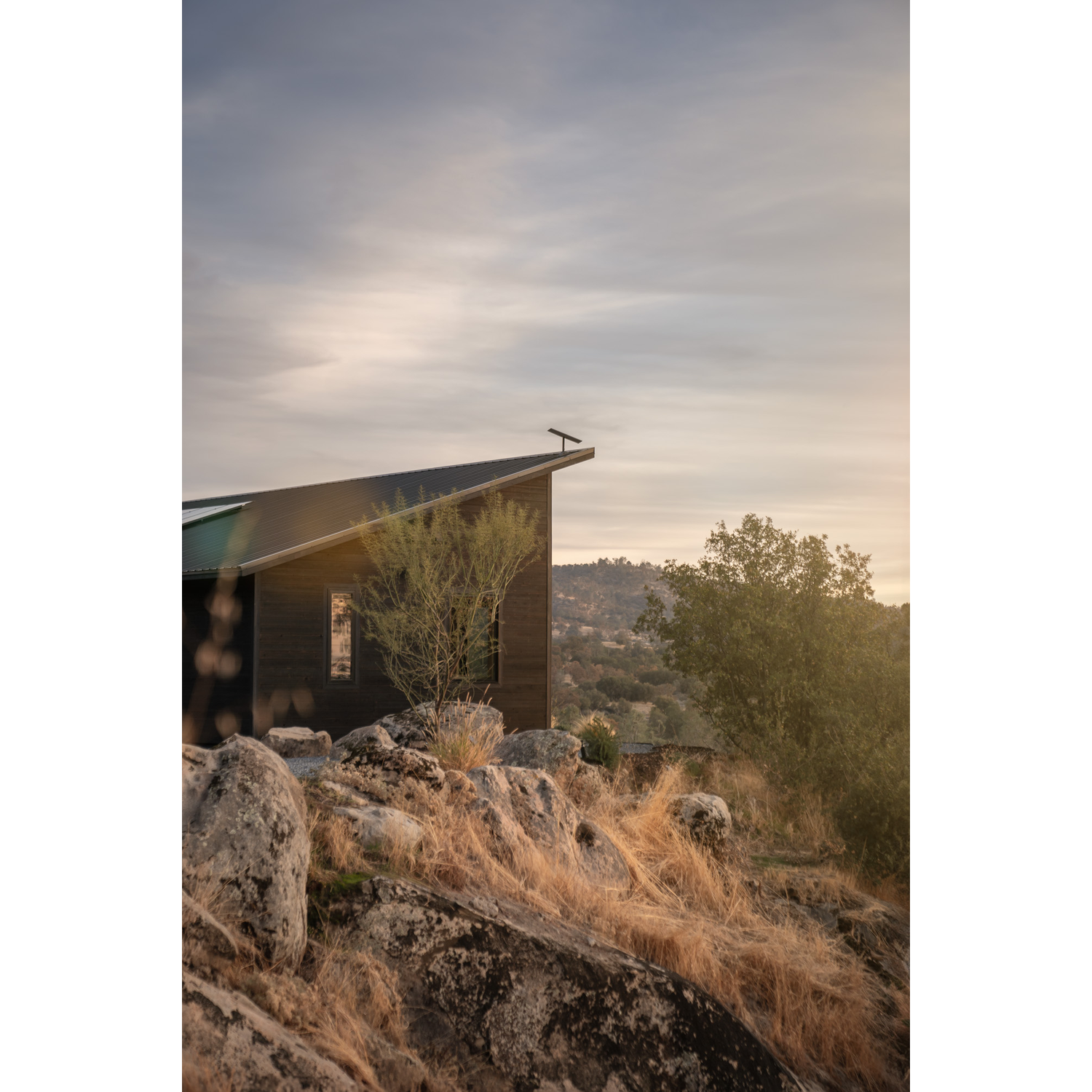 Een modern donker huis op een rotsachtige heuvel bij zonsondergang met droge grasvegetatie.