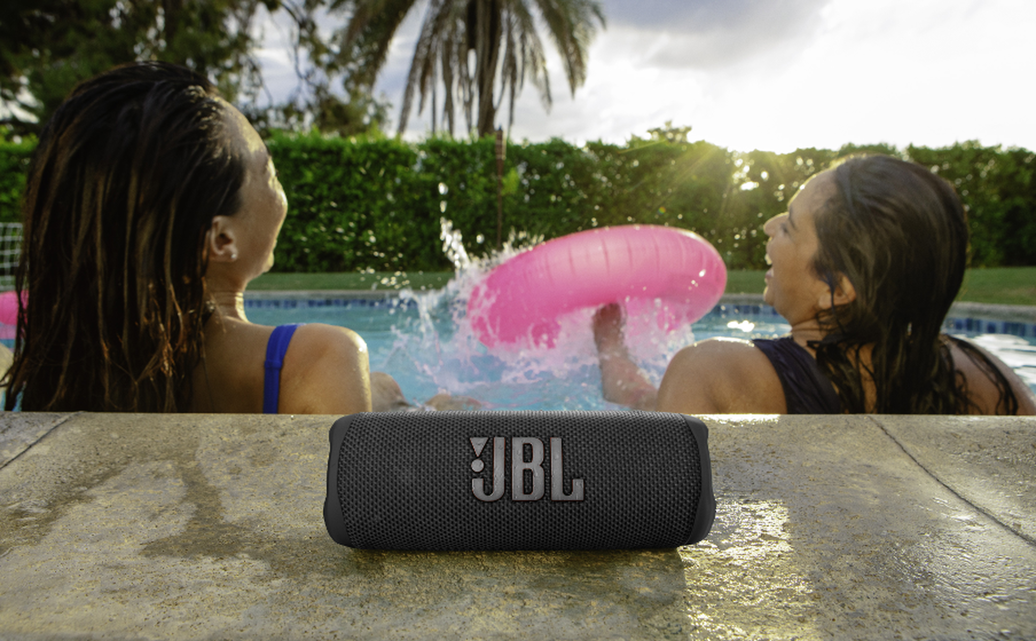 Un altavoz JBL negro descansa en el borde de piedra. Dos mujeres chapotean en una piscina.