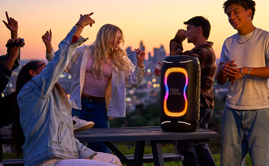Des amis dansent autour d'une enceinte JBL avec des lumières au coucher du soleil.