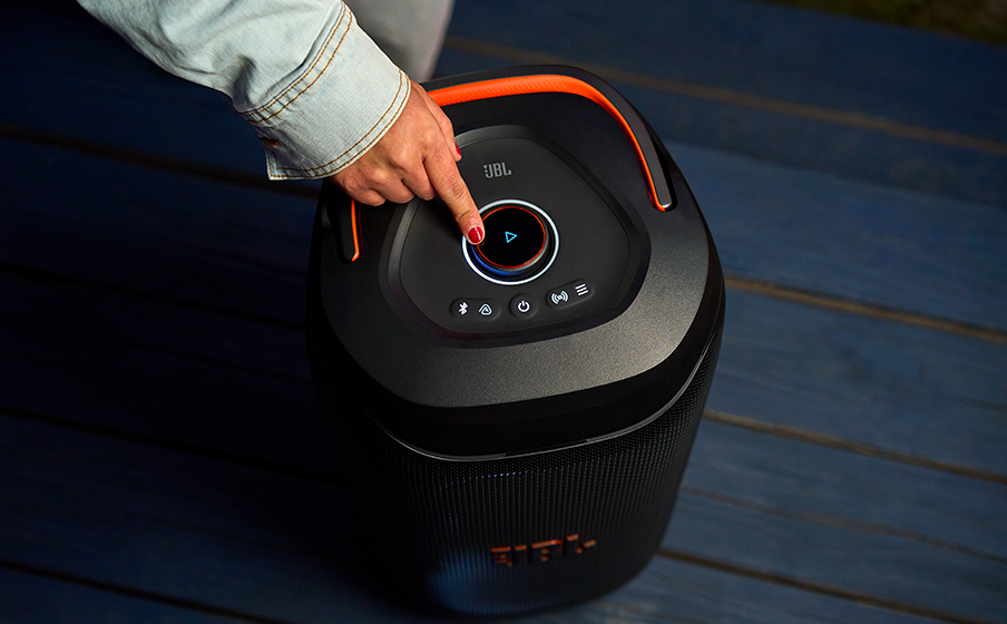 Main appuie sur play sur enceinte JBL avec accents orange sur bois bleu.