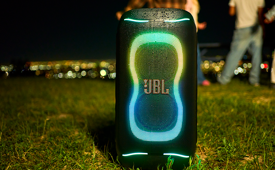 Un haut-parleur JBL éclairé sur l'herbe la nuit, avec des lumières de ville floues à l'arrière-plan.