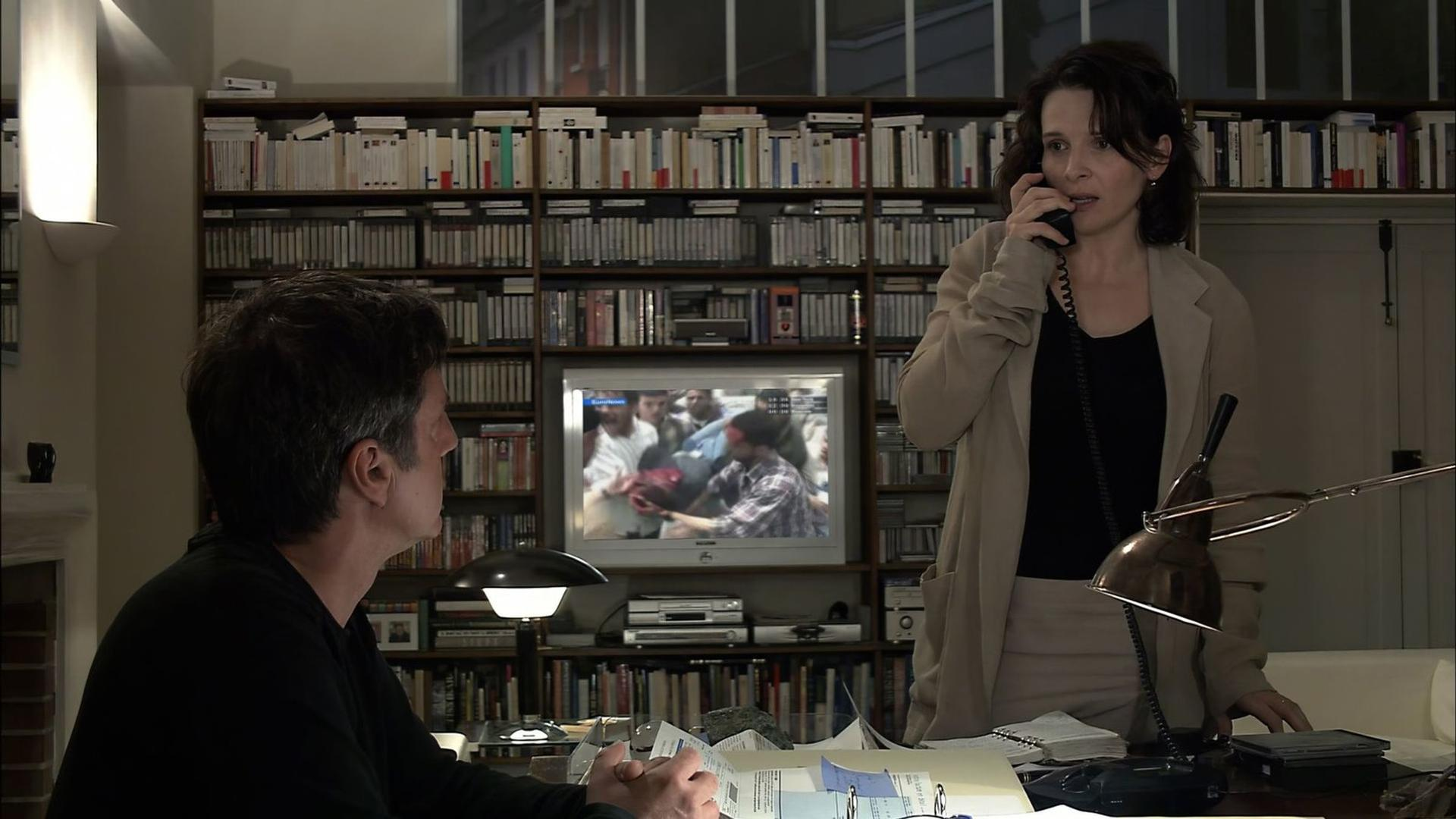 Eine Frau am Telefon steht an einem Schreibtisch, Blick auf ein Bücherregal. Ein Fernseher zeigt eine Menge.