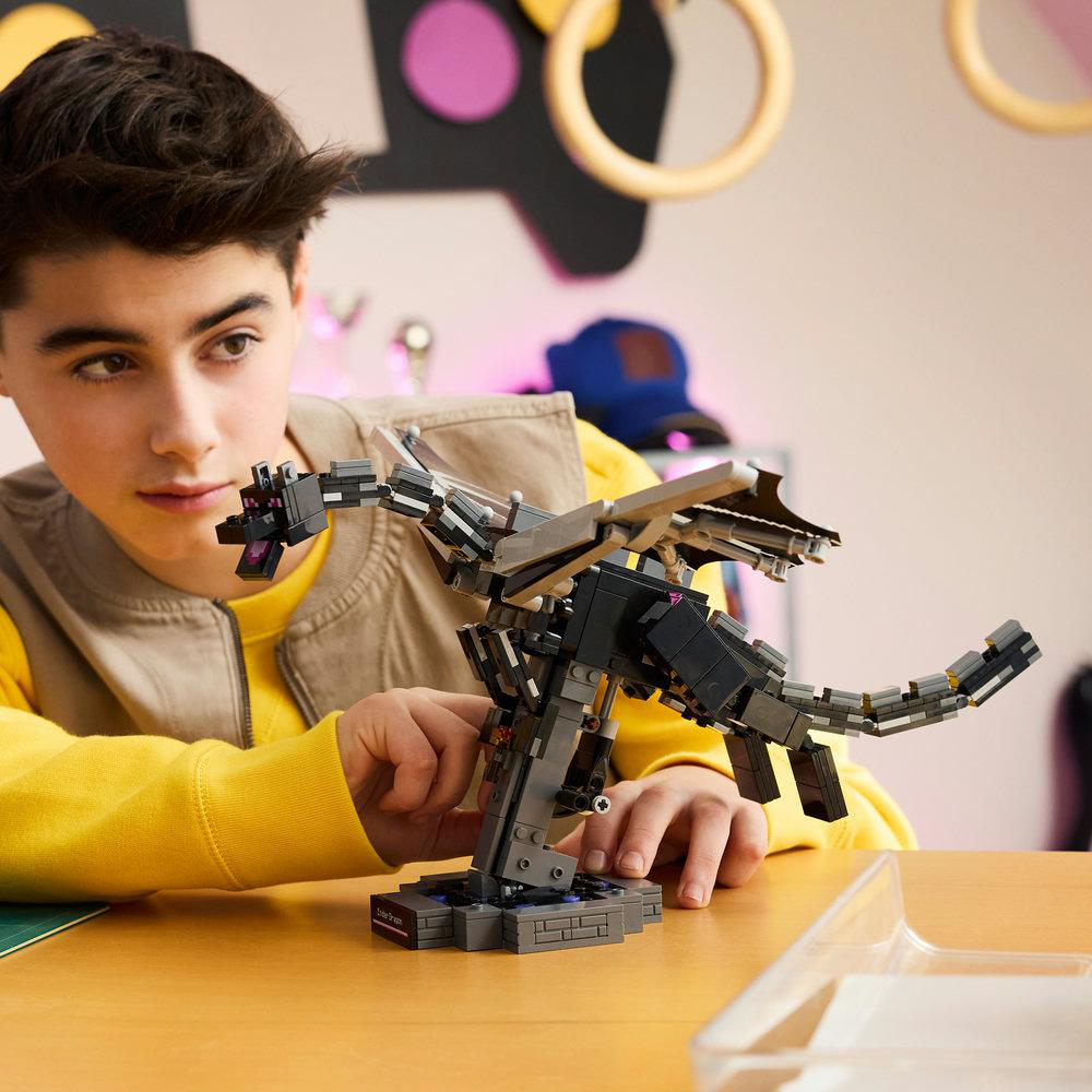 Ein Junge baut einen grauen Lego-Drachen auf einem Holztisch. Er trägt ein gelbes Sweatshirt.