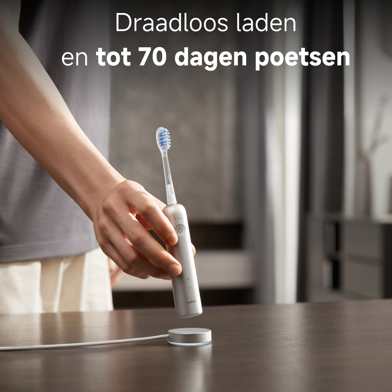 A hand holds a silver toothbrush over a charging base.