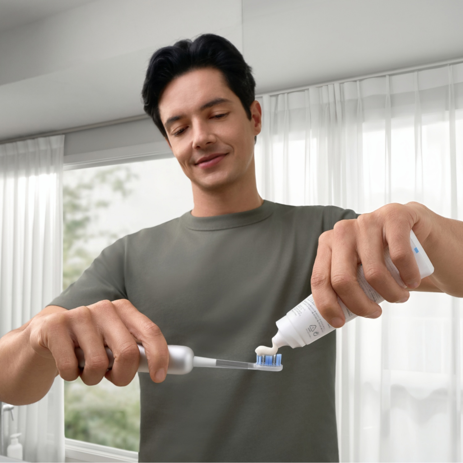 A man in a green shirt smiles while applying white toothpaste onto a white electric toothbrush in a bright bathroom.