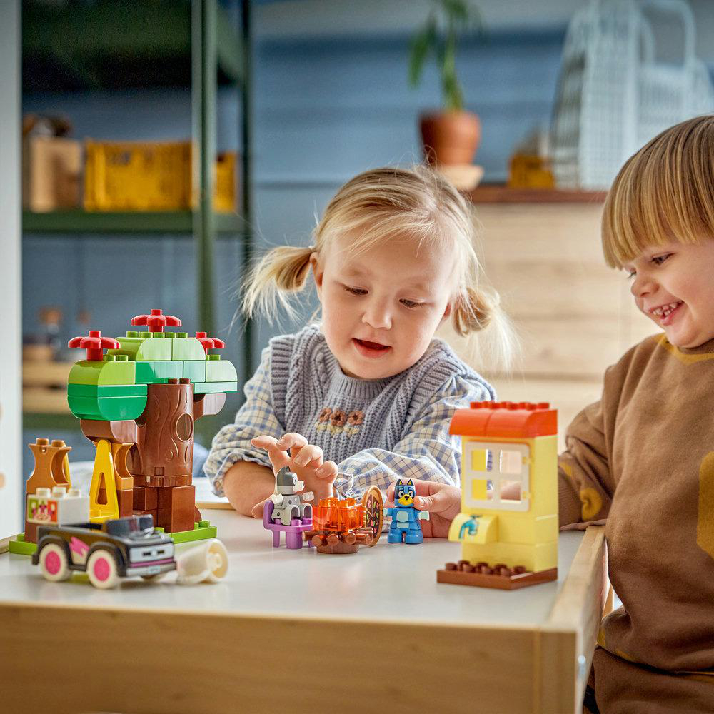 Zwei Kinder spielen mit bunten Bausteinen, darunter ein Baum, ein Auto und Figuren.