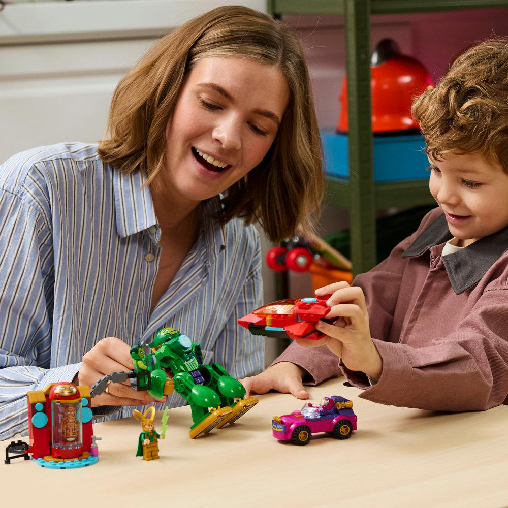 Frau und Kind spielen mit bunten Lego-Spielzeugen: ein grüner Roboter, ein rotes Raumschiff, ein rosa Auto, Figuren.