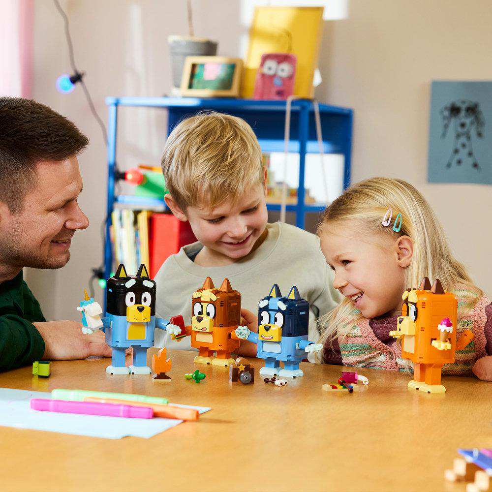 Vater und Kinder bauen mit Bluey-Spielzeugfiguren an einem Holztisch.