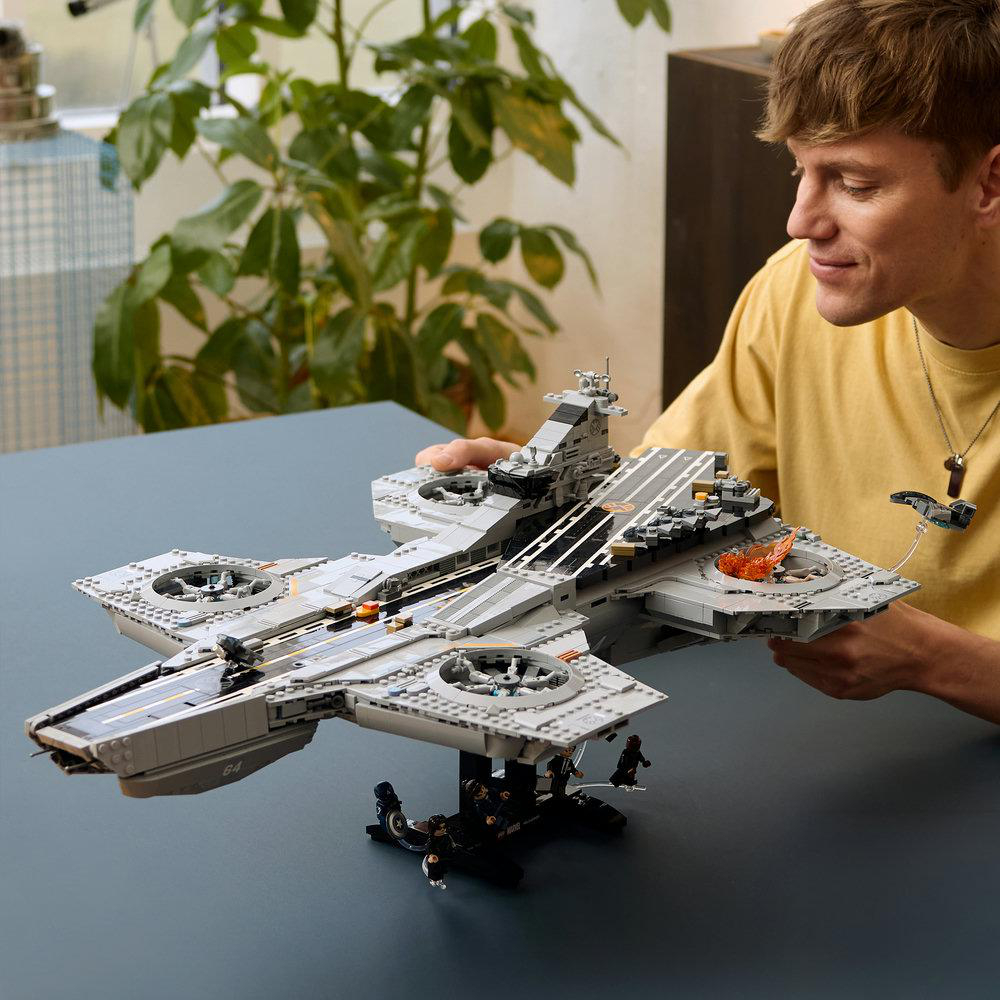 Mann in gelbem Hemd hält ein graues Lego Quinjet-Flugzeugmodell mit orangen Flammen.