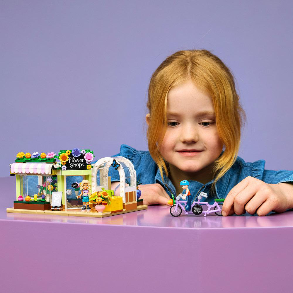Mädchen mit roten Haaren spielt mit einem Lego-Blumenladen und Fahrradset auf lila Tisch.