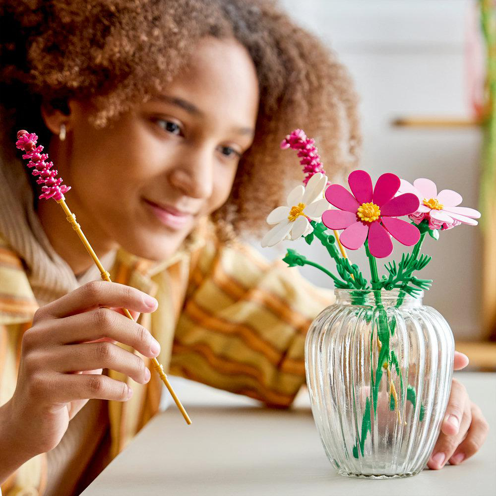 Ein Mädchen mit Locken hält eine gelbe Blume vor eine Glasvase mit bunten Blumen.