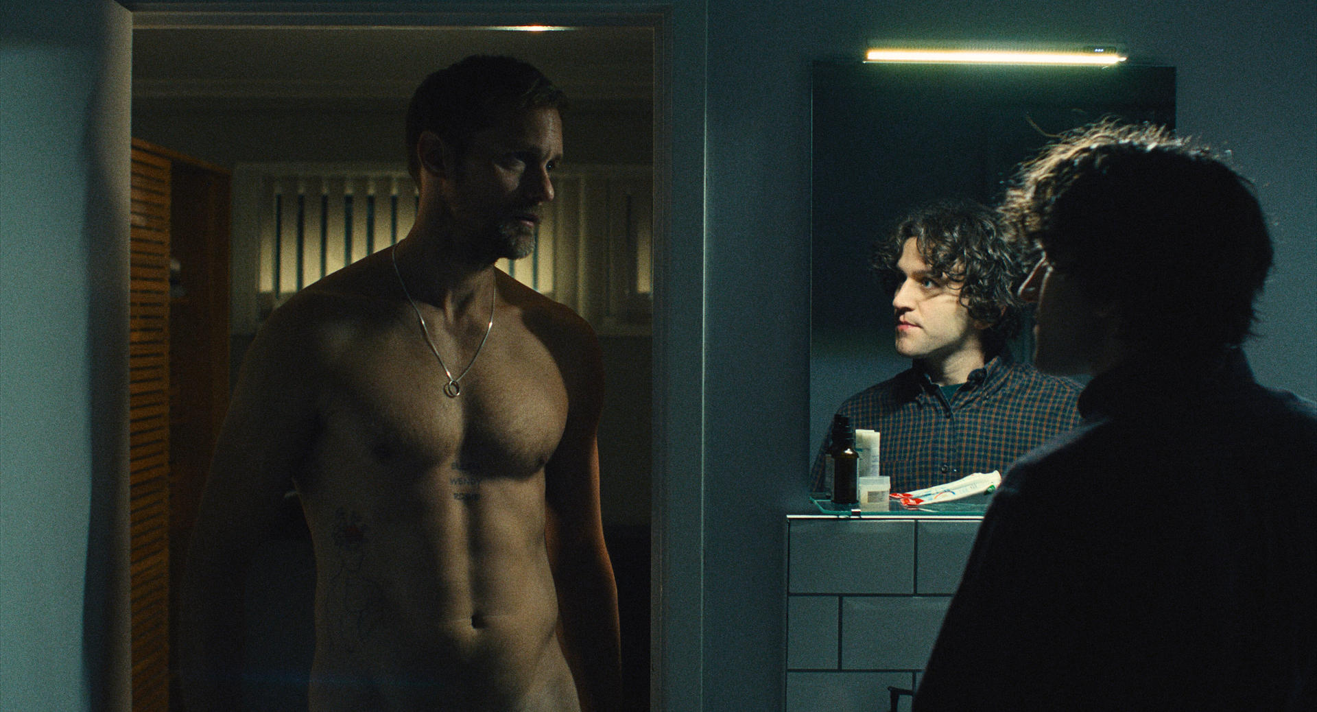 Ein Mann mit Kette steht vor einem Spiegel, ein anderer Mann blickt hinein.