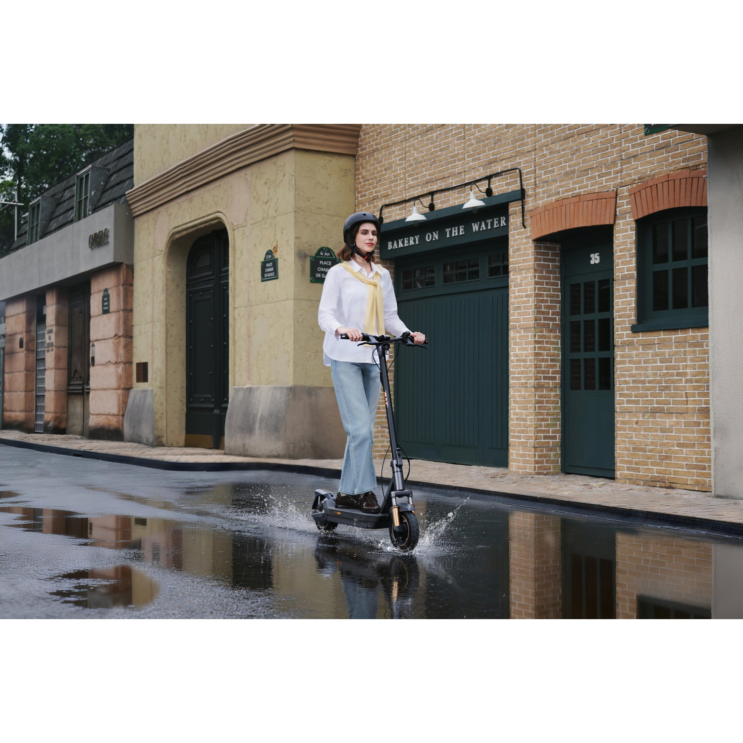 Een vrouw rijdt met een elektrische scooter door een plas voor een bakstenen gebouw.