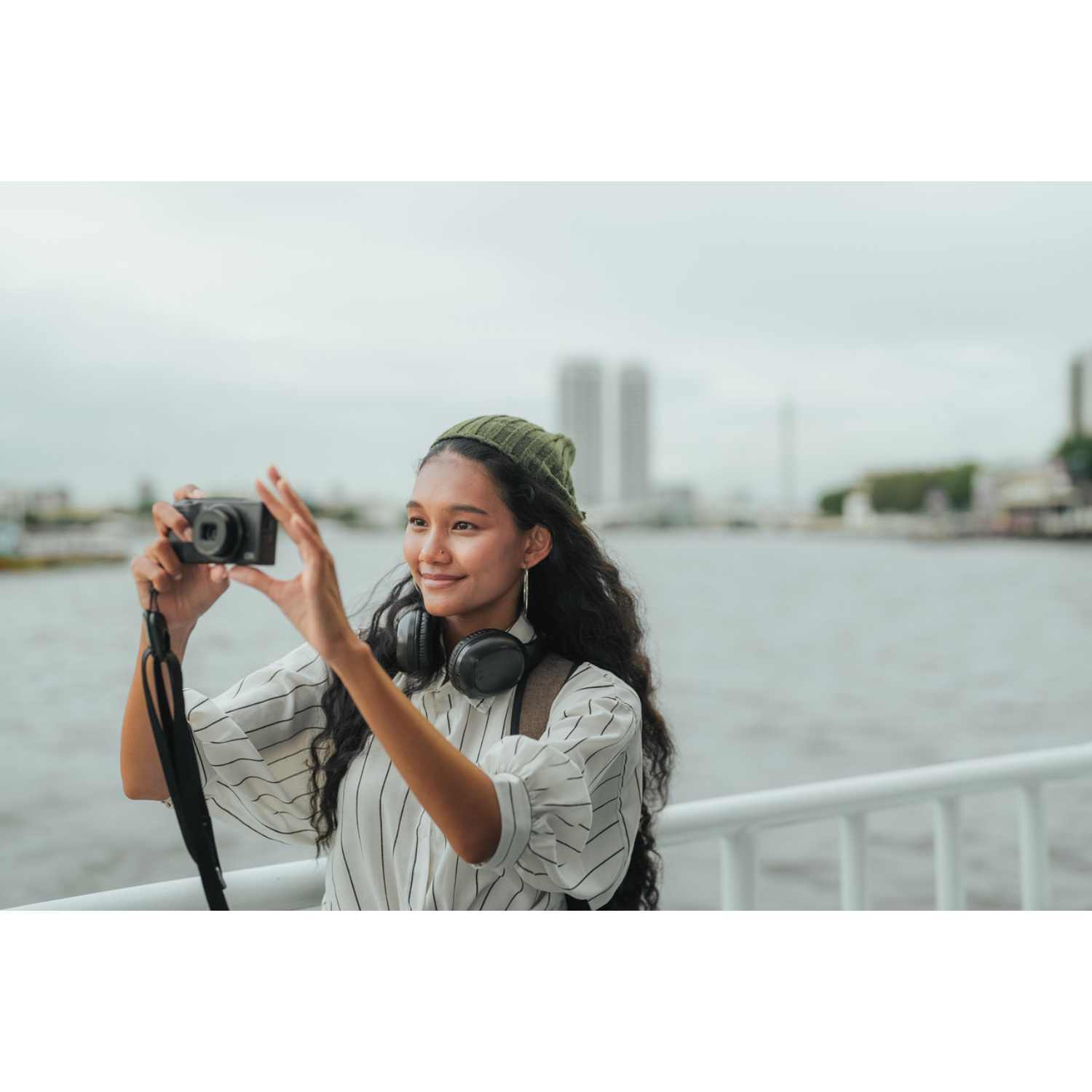 Una joven con auriculares y gorro verde toma una foto con una cámara junto al agua.