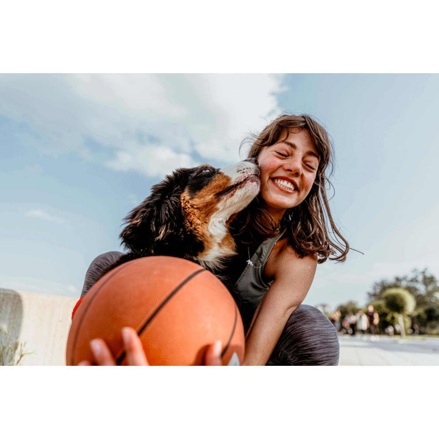 Un perro besa a una mujer riendo con una pelota de baloncesto bajo el cielo azul.
