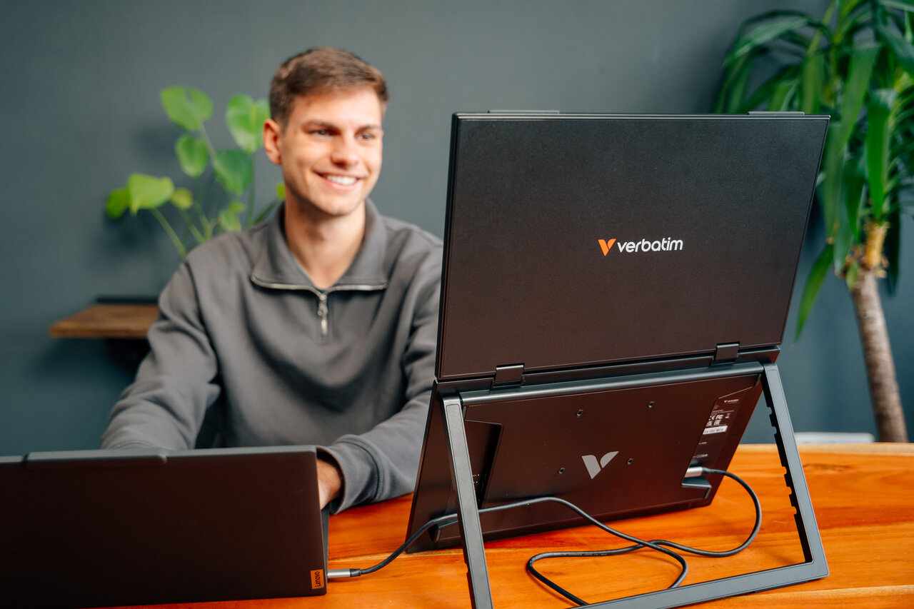 Ein Mann sitzt an einem Holztisch und arbeitet an zwei Laptops, einer davon ist aufgeklappt.