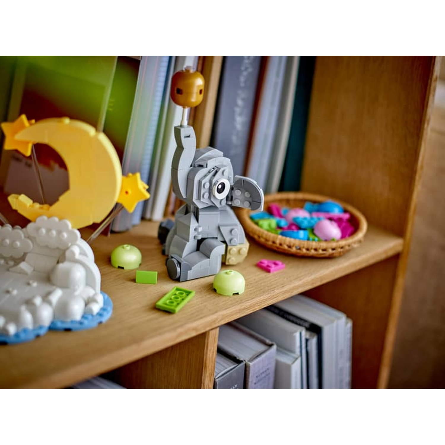 Grijze Lego-olifant zit op een plank naast een gele maan en een kom met kleurrijke Lego-onderdelen.