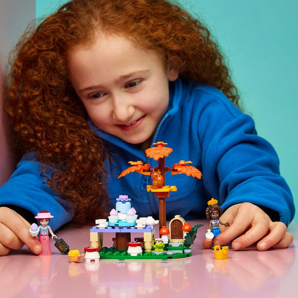 Ein Mädchen mit lockigem rotem Haar lächelt und spielt mit einem Lego-Set aus Kuchen, Baum und Figuren.