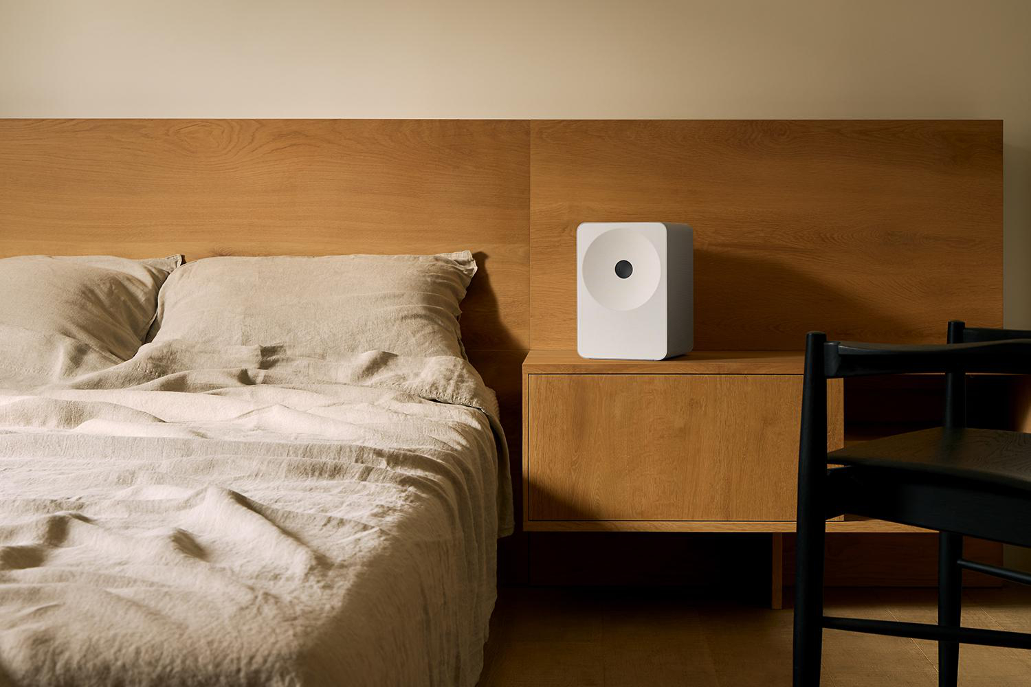 Ein weißer Lautsprecher auf einem Nachttisch neben einem Bett mit Holzkopfteil.