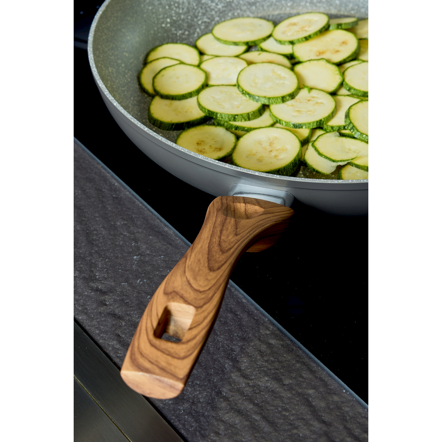 Zucchine affettate in padella con manico in legno su piano cottura nero.