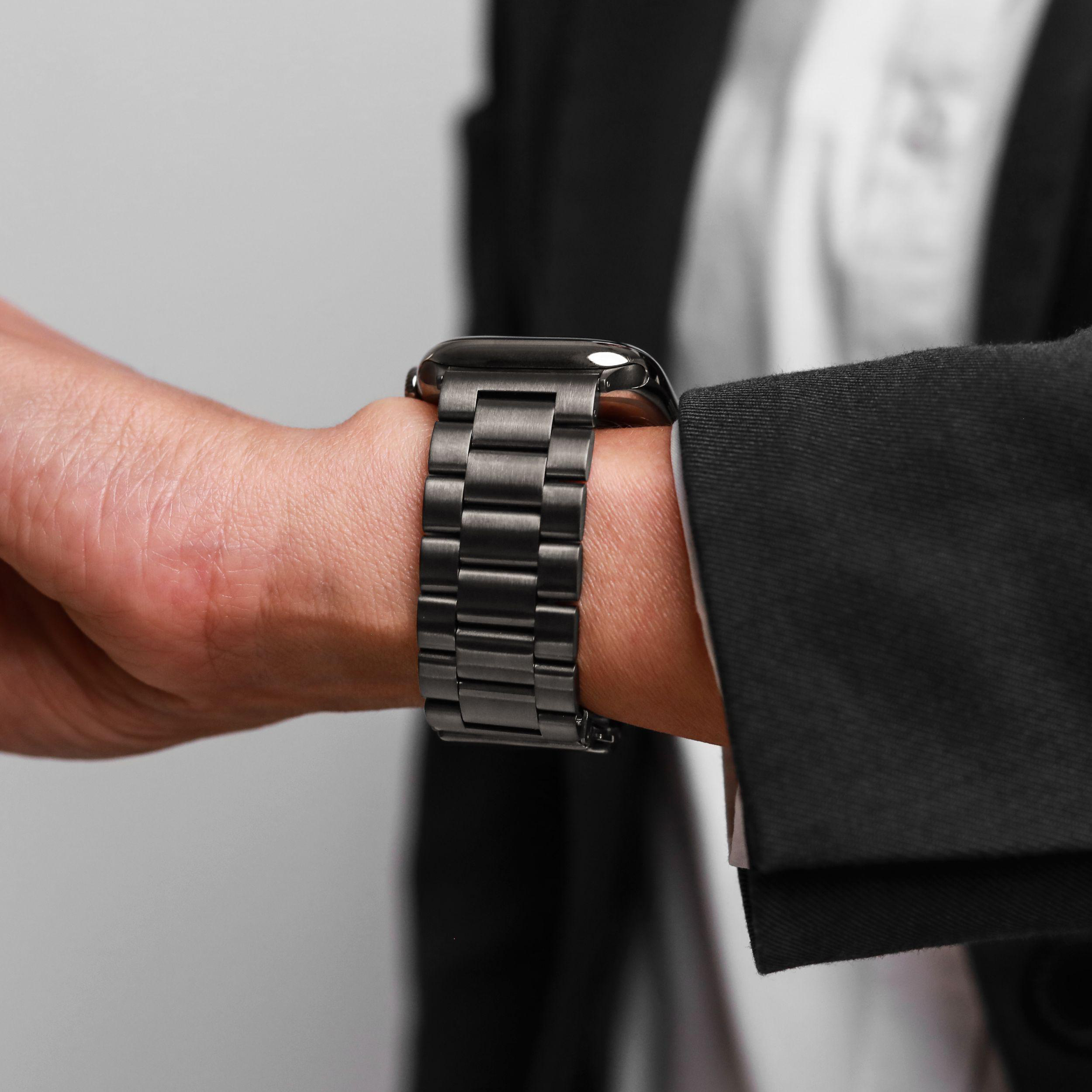 Eine dunkle Uhr mit Metallarmband an einem Handgelenk, das einen schwarzen Anzug trägt.