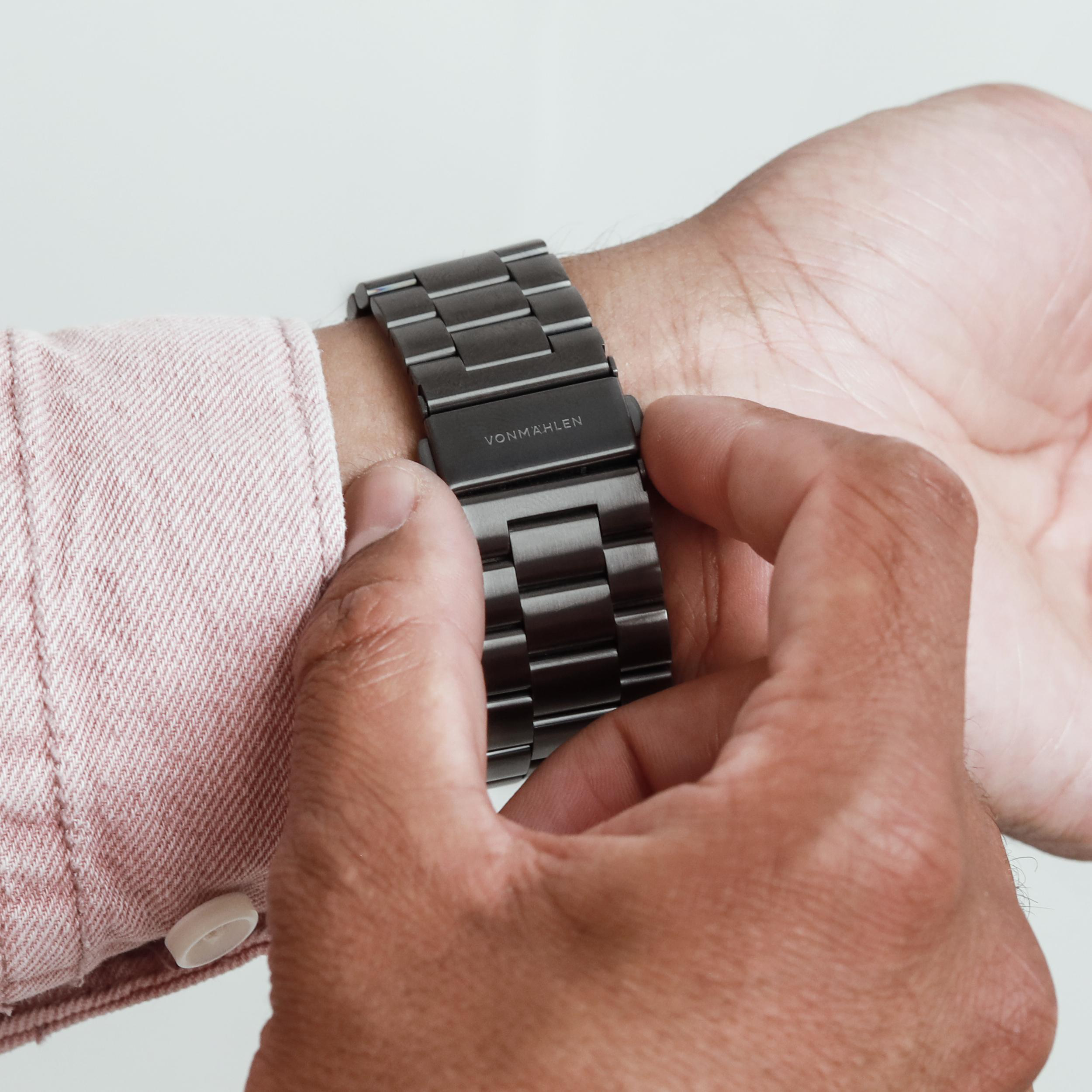 Mann schließt eine dunkle Uhr mit Metallarmband an einem rosa Hemd.