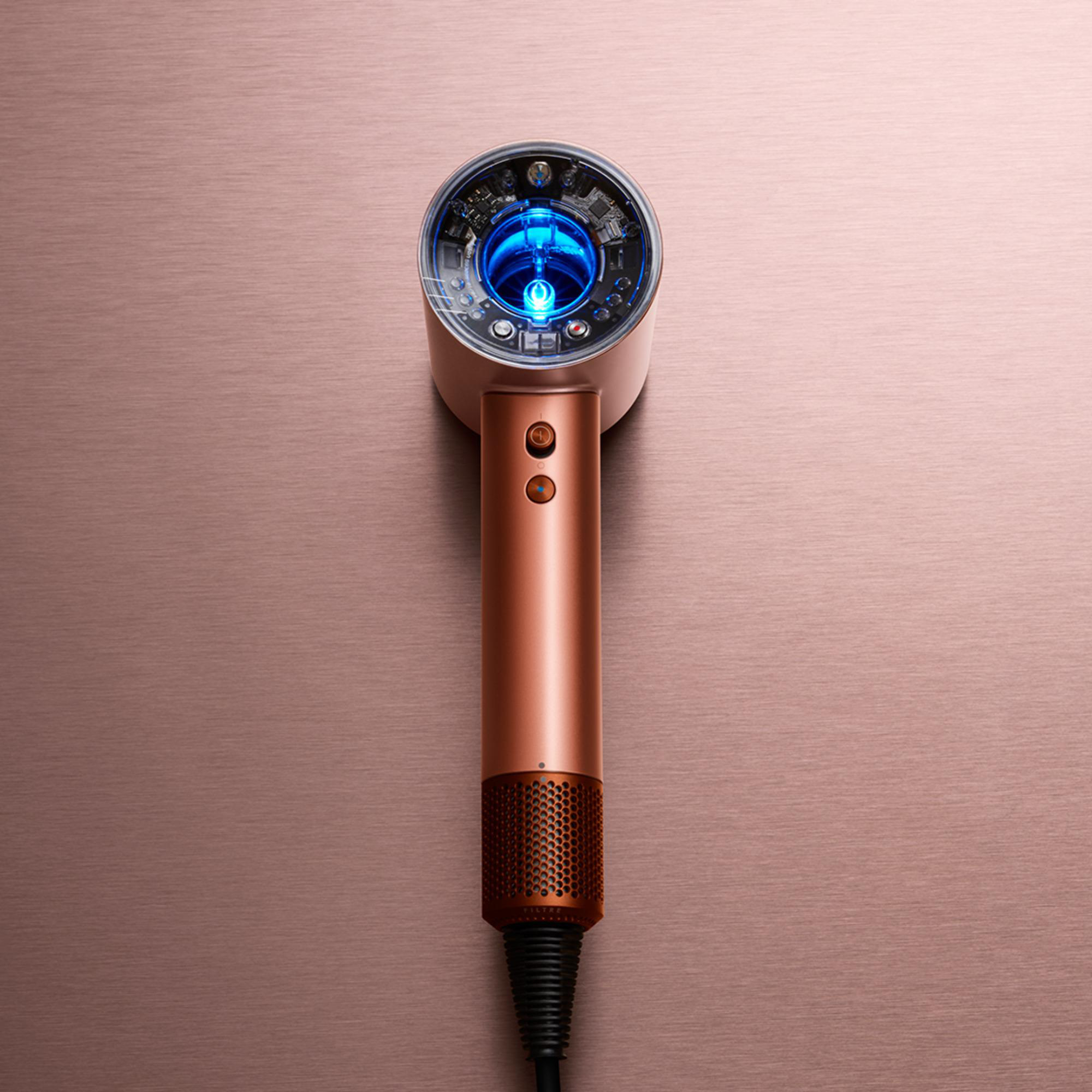 Ein roségoldener Haartrockner mit blauem Licht im Inneren, auf einem metallischen Hintergrund.