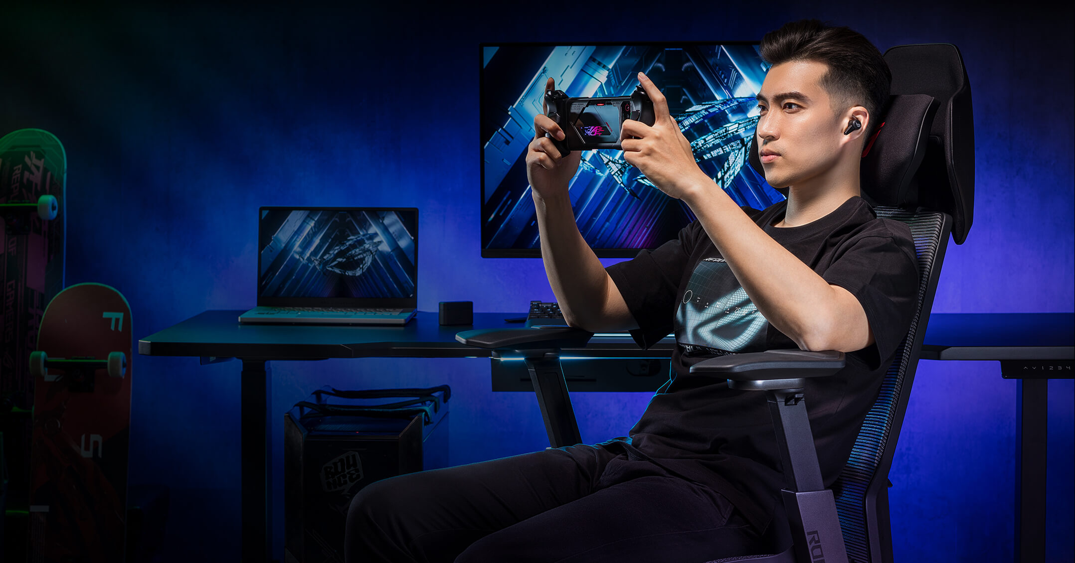 Ein Mann sitzt in einem Gaming-Stuhl und spielt auf einem Handheld-Gerät. Ein Laptop und ein Monitor sind zu sehen.