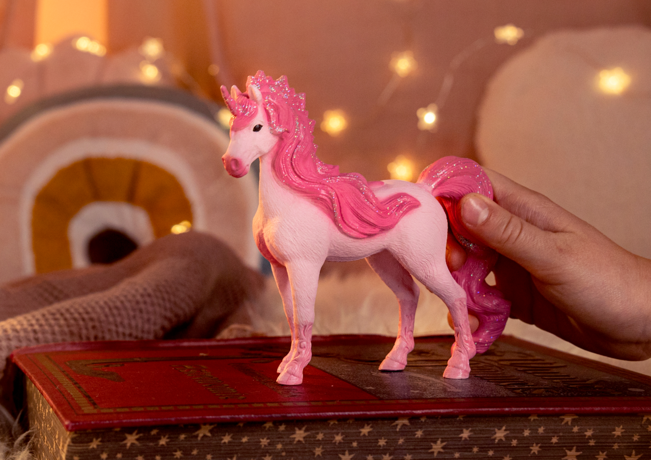 Ein rosa Glitzer-Einhorn-Spielzeug steht auf einem Buch. Eine Hand hält seinen Schwanz.