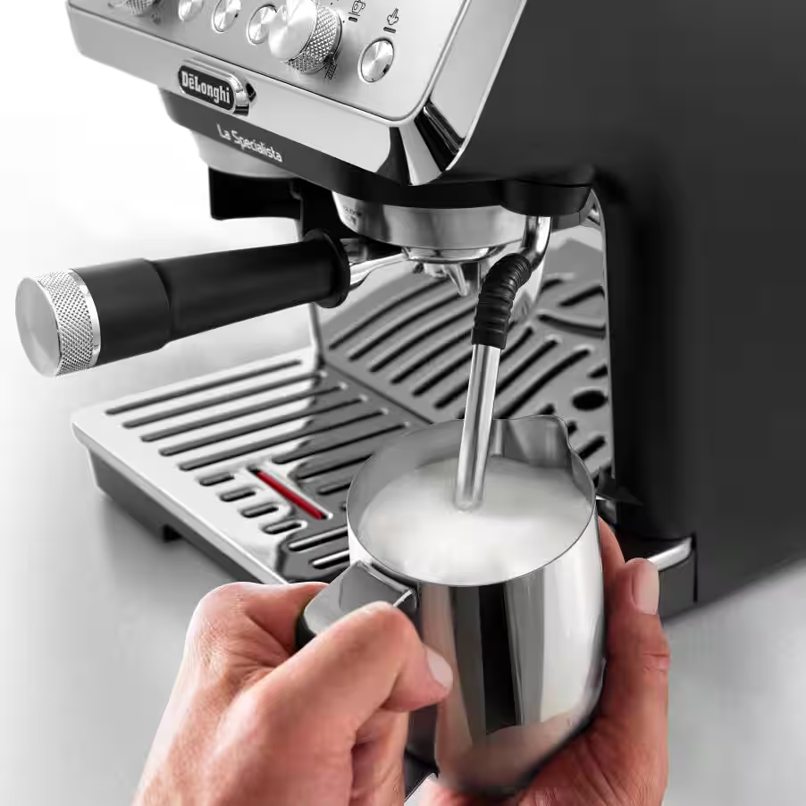 Una mano espumando leche en una jarra metálica junto a una máquina de espresso.