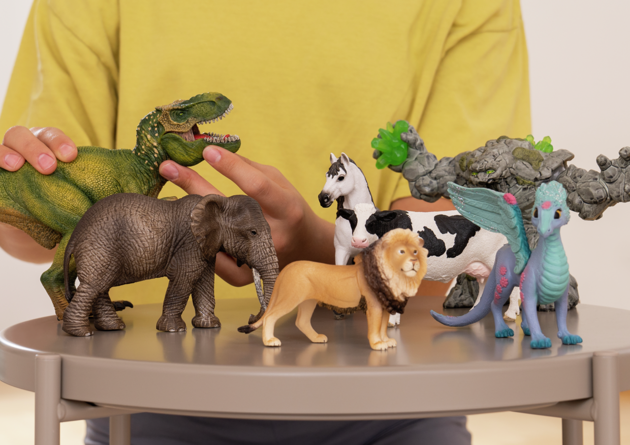 Eine Person hält eine grüne Dinosaurierfigur über einen Elefanten, Löwen, Pferd, Kuh und Drachen.