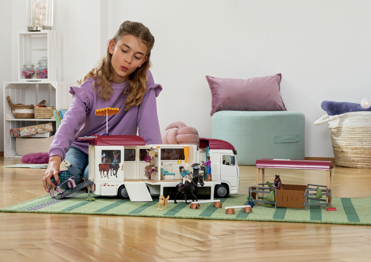 Mädchen spielt mit Spielzeug-Pferdeanhänger, Stall und Figuren auf grünem Teppich.