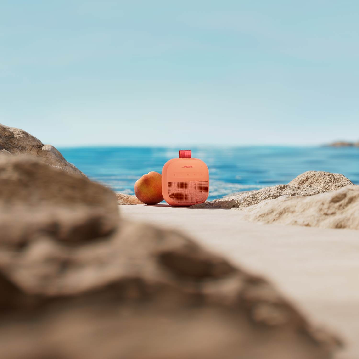 Een oranje Bose speaker en een perzik op het strand met de zee erachter.