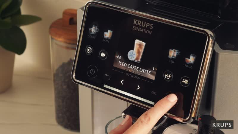 Ein Finger drückt auf den OK-Knopf des Touchscreens einer Kaffeemaschine, die "ICED CAFFE LATTE" anzeigt.