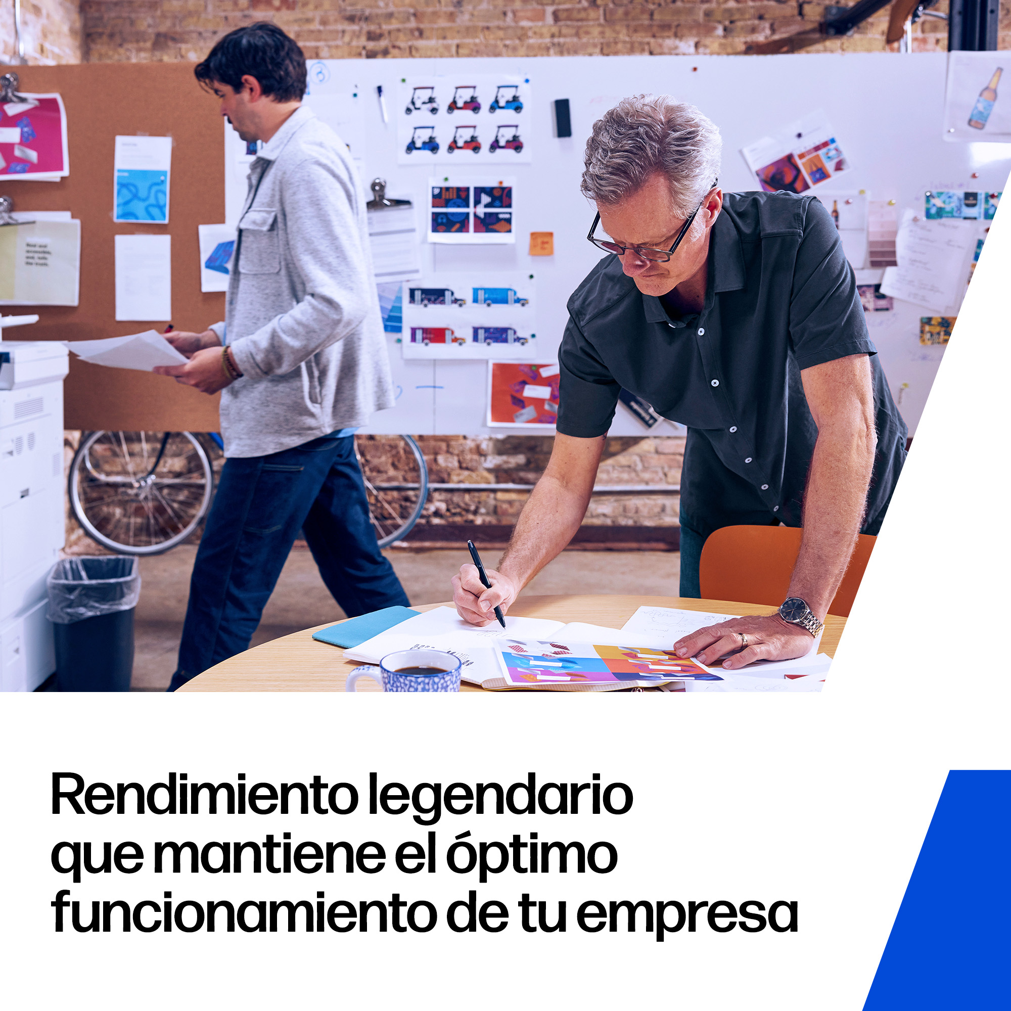 Dos hombres trabajan en una oficina. Uno está junto a un tablón de anuncios, el otro sentado.