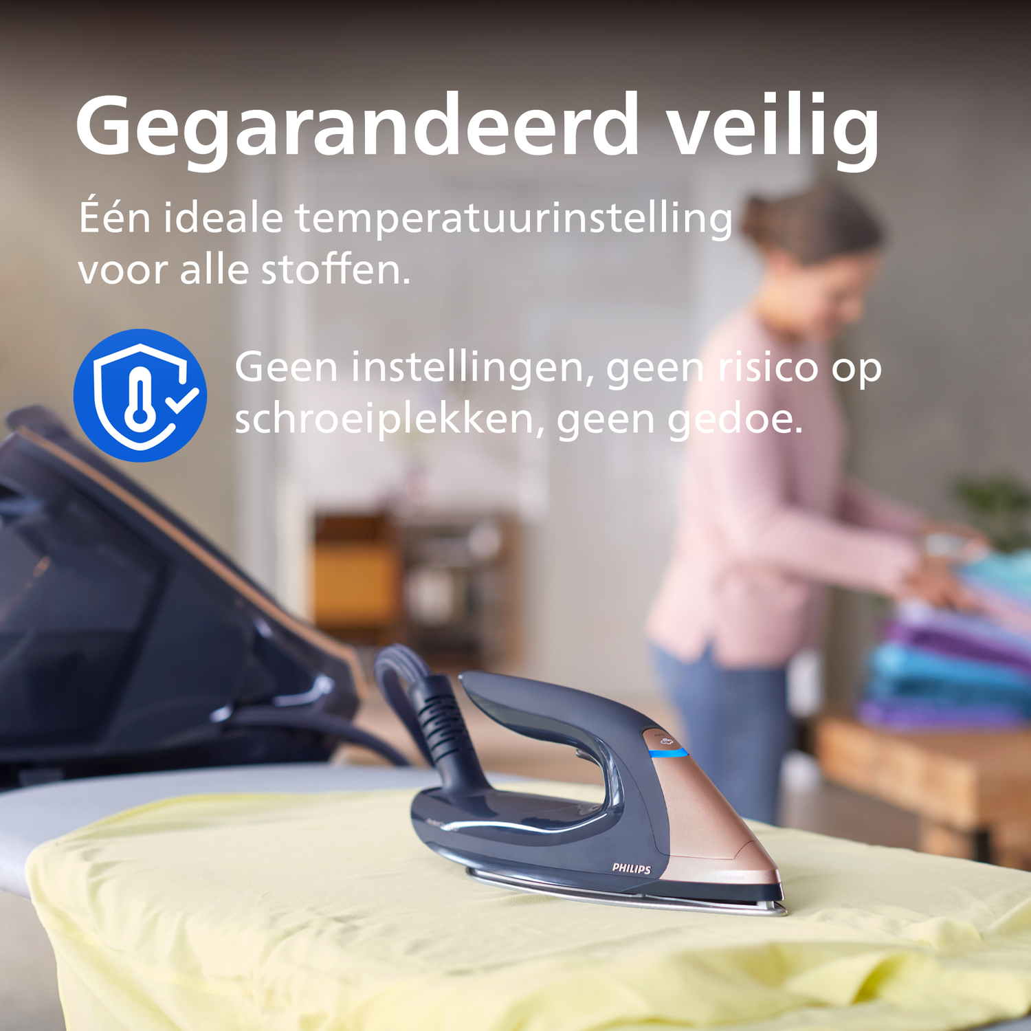Een strijkijzer met veiligheidssymbool op een strijkplank. Een vrouw vouwt kleding op de achtergrond.