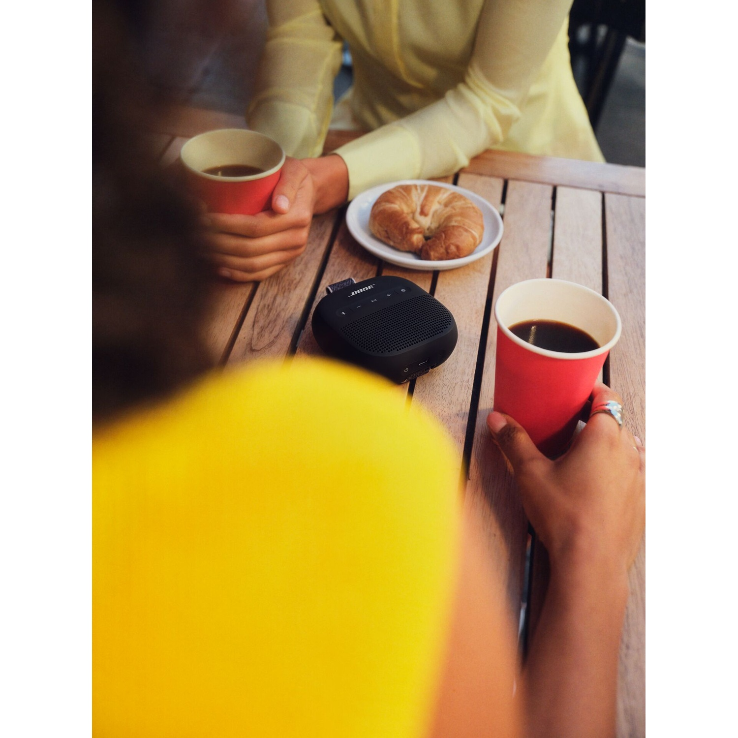 Twee mensen drinken koffie aan een houten tafel met een croissant en een luidspreker.